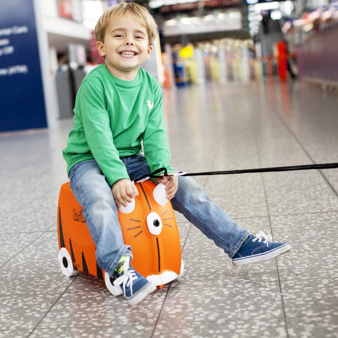 Kind sitzt lachend auf orangefarbenem Kinderkoffer in Tierform zum Fahren und Ziehen im Flughafen