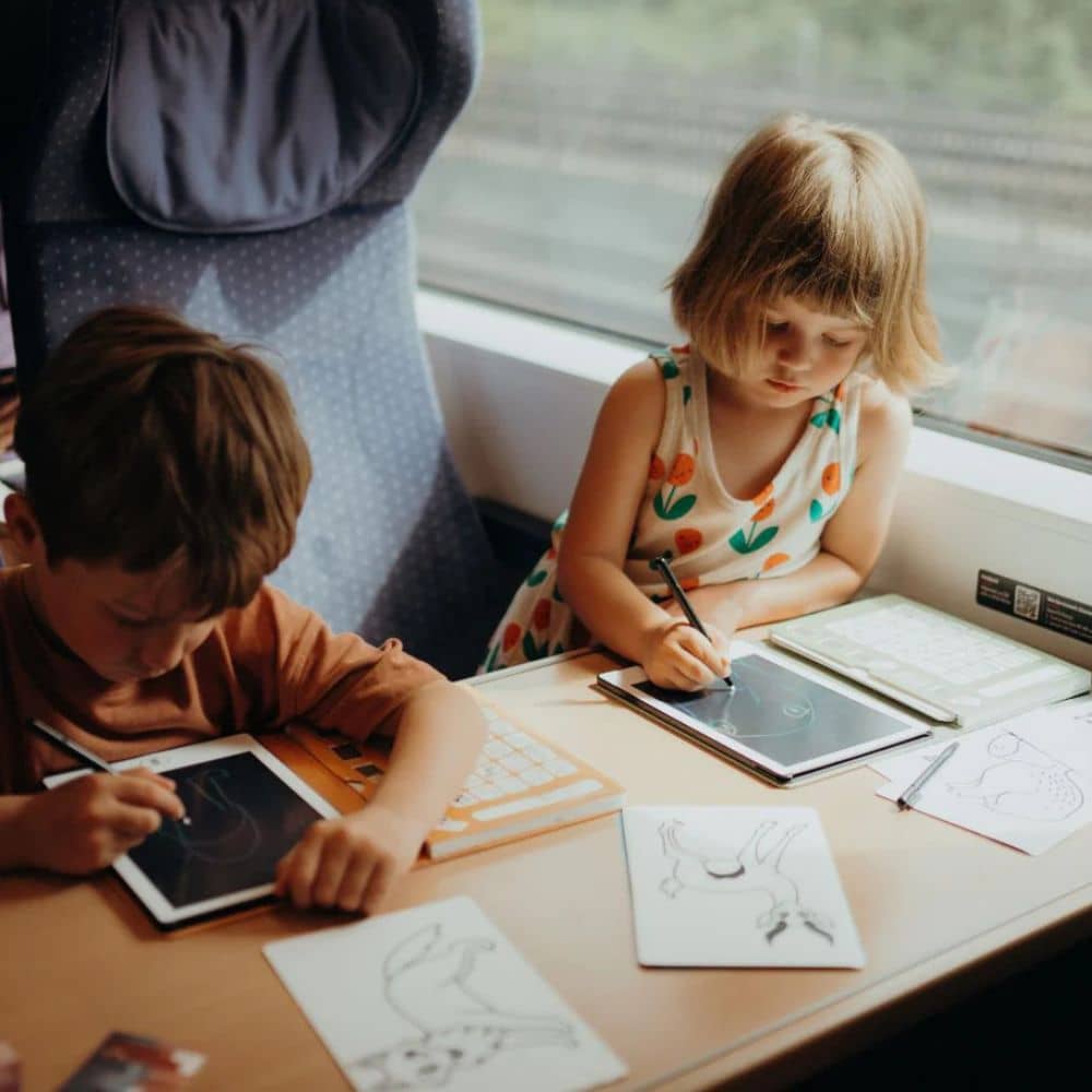 Zwei Kinder beschäftigen sich mit kreativem Spielzeug während einer Zugfahrt mit Tablets und Malsets – Spiele für unterwegs kaufen