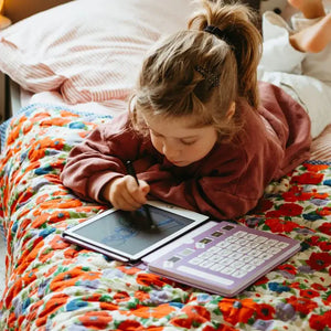 Mädchen zeichnet mit Grafiktablett Kidydraw Mini – Prinzessinnen auf einem bunten Bett mit Blumenmuster.
