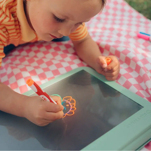 Kind malt mit bunten Stiften auf der Leuchttafel Kidydraw Pro auf einer Decke im Freien
