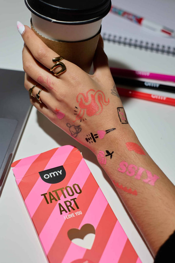 OMY Tattoo Marker Set für Kinder mit bunten Designs auf der Hand und Kaffeebecher im Hintergrund