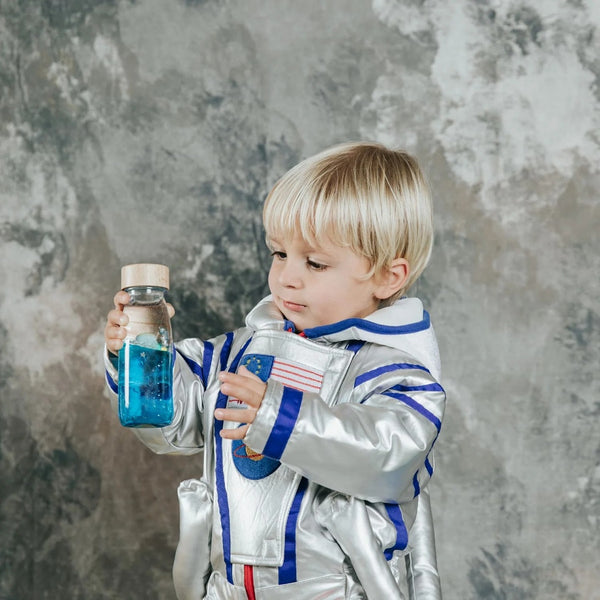 Kleines Kind im Raumanzug mit einer Sensorikflasche für Kinder in der Hand, die glitzernde Flüssigkeit enthält